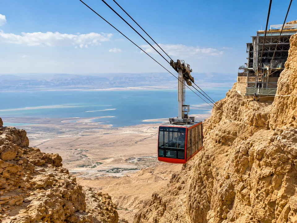 A red cable car cabin rising up a desert cliff, the Dead Sea plain far below