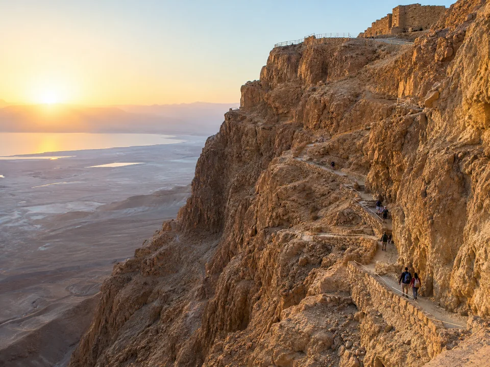 A zigzag path climbing a steep desert cliff in early light, with tiny hikers seen from behind