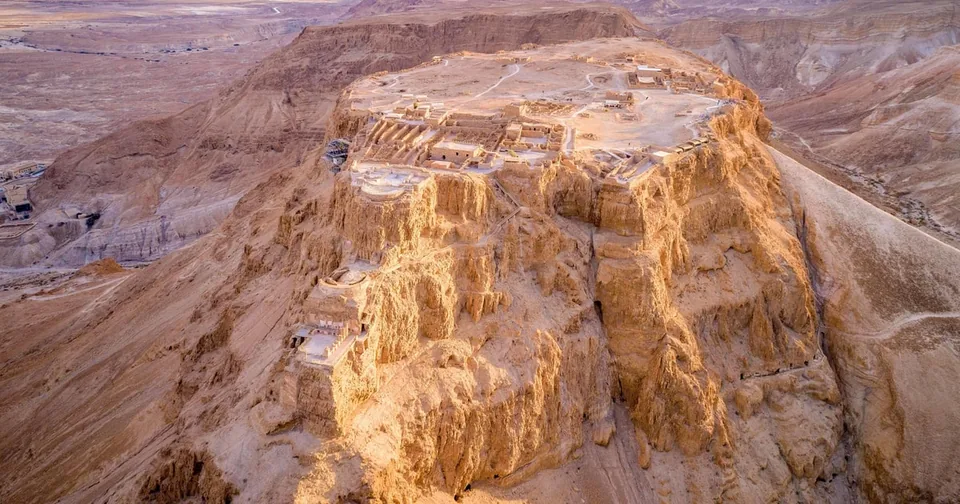 The flat summit of a desert mesa from the air, ruins on top and cliffs falling away