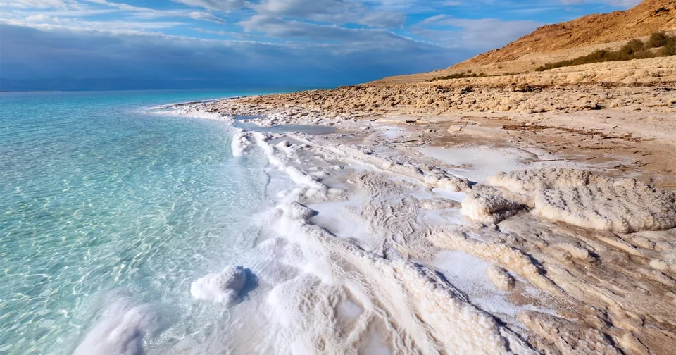 A white salt-crusted shore curving along turquoise water under desert hills